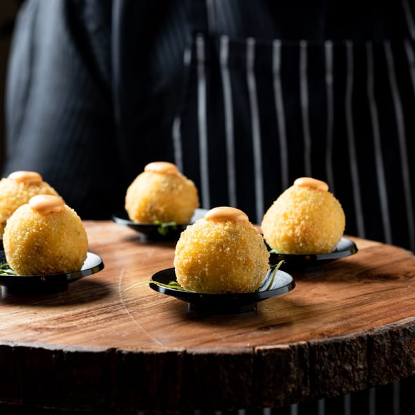 Five golden-fried arancini balls with sauce on black plates, served on a rustic wooden board.