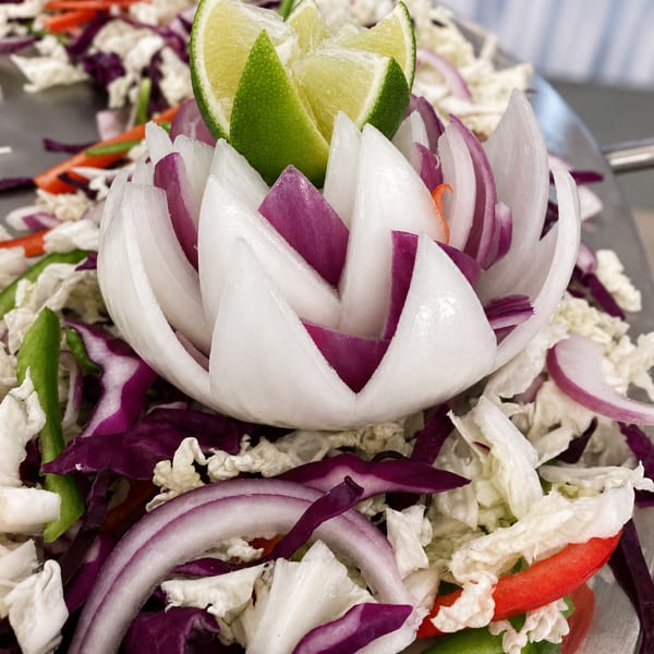 A carved onion flower with lime wedges, surrounded by shredded cabbage, red onion, and bell peppers.