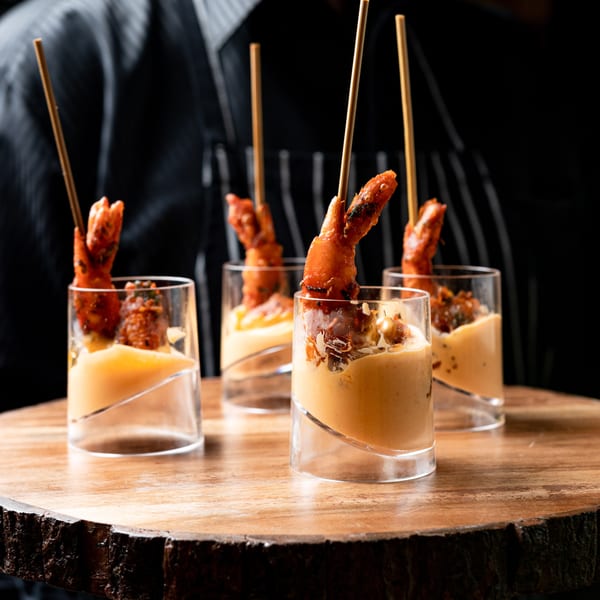 Four shrimp skewers in small glasses with sauce, held on a rustic wooden tray.