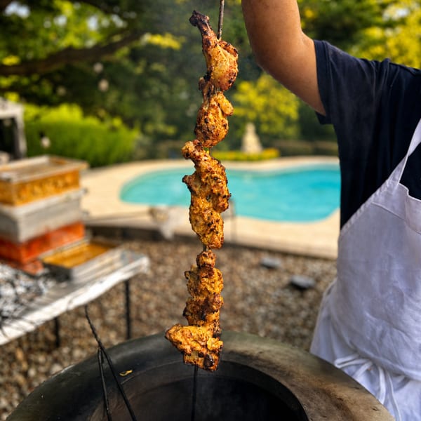 A chef lowers chicken tikka skewers into a tandoor oven beside a swimming pool.