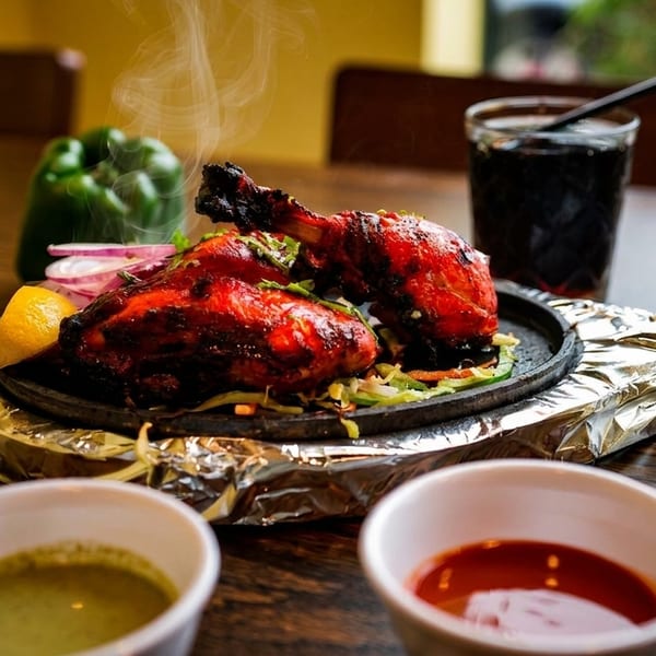 Steaming tandoori chicken on a sizzling platter with lemon, onions, sauces, and a drink.