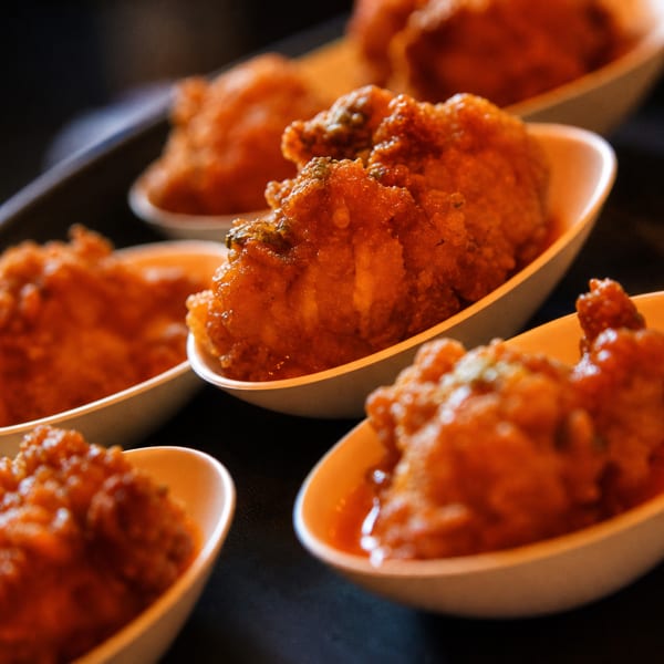 Close-up of several small white dishes filled with crispy, orange-glazed fried chicken bites on a dark tray.