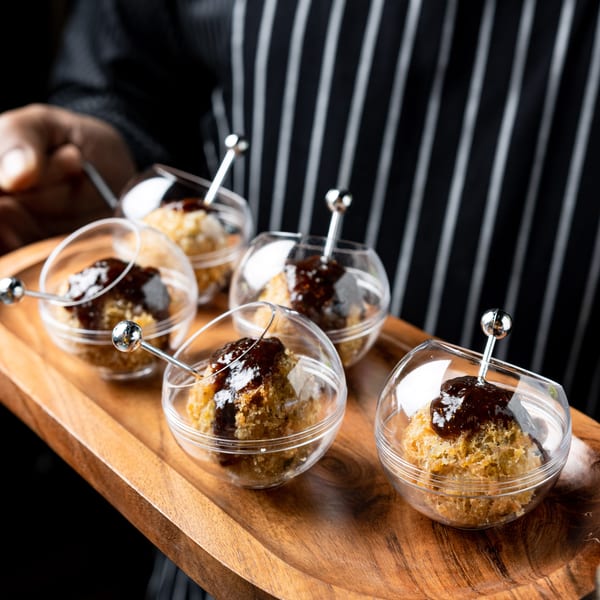 A server in a striped apron holds a wooden tray with six individual dessert cups, each with a pastry and dark sauce.