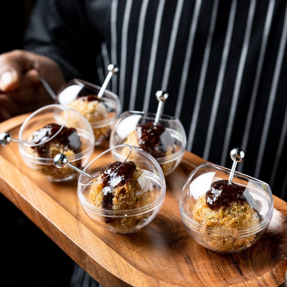 A server in a striped apron holds a wooden tray with six individual dessert cups, each with a pastry and dark sauce.