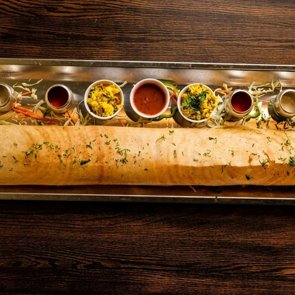 A long dosa with various chutneys and sambar in small bowls on a metal tray.