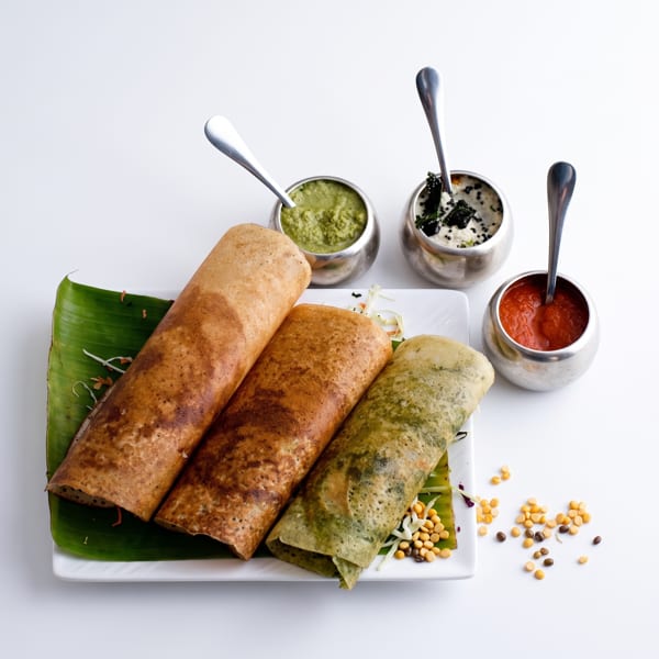 Three dosas, two brown and one green, on a banana leaf with three chutneys in steel bowls.